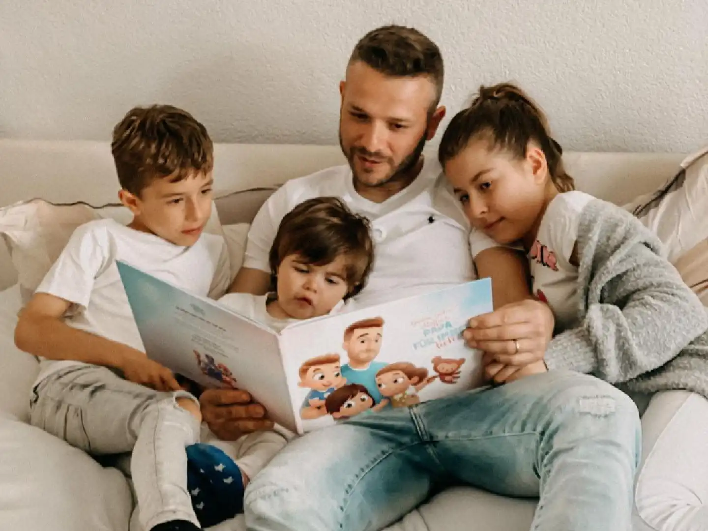 Der Vater hält seine drei Kinder im Arm und liest ihnen ein Buch mit lustigen Geschichten über sie vor.